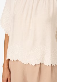 Blusa bianca con tessuto testurizzato e ricami floreali sull'orlo. Le maniche sono ampie e fluide. Abbinata a un pantalone beige.