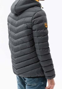 Chaqueta gris acolchada con capucha, que presenta un acolchado en forma de chevron, detalles amarillos en los hombros y un parche circular en el brazo izquierdo.