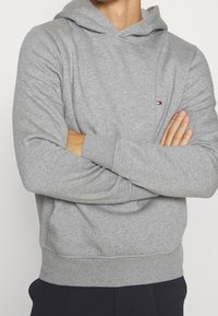 Sudadera gris con capucha, hecha de material suave. Presenta puños y dobladillo de canalé, un pequeño logo en el pecho y un corte relajado.