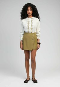 Blouse blanche avec broderie dorée, manches longues et fermeture à boutons ; jupe portefeuille olive avec boutons sur le devant ; sandales noires à bout ouvert.