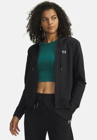 Felpa nera con cerniera e cappuccio, dotata di tasche frontali e dettagli in argento. Il modello indossa un top corto verde a coste sotto.