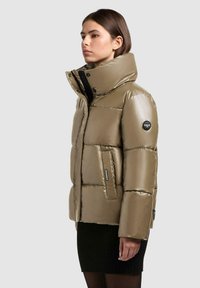 Giacca puffer khaki con colletto alto, finitura lucida, dettagli cuciti e una patch con logo sulla manica; presenta tasche laterali.