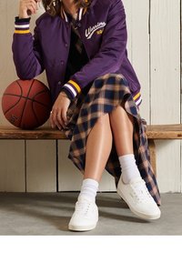 Osoba sedící na dřevěné lavičce, oblečená v fialové varsity bundě, kostkované sukni, bílých teniskách a držící basketbal pod jednou paží.