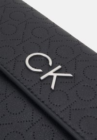Superficie in pelle nera testurizzata con motivi a cuore in rilievo e un logo "CK" metallico argentato su una patta.