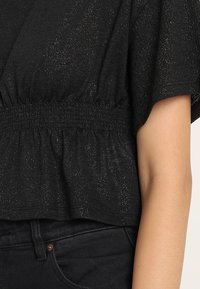 Sort top med en tekstureret overflade, der har en smocket talje og korte, brede ærmer. Stoffet inkluderer subtile metaltråde.