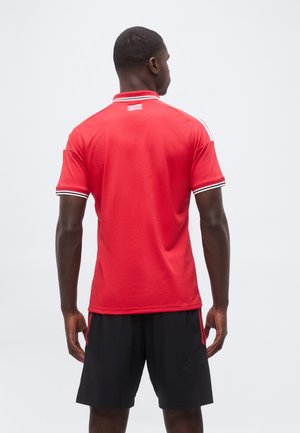 Homme portant un t-shirt rouge à manches courtes avec des bandes blanches aux épaules et un short noir, debout dos à la caméra sur un fond uni.