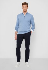 Helles blaues Zip-Hoodie über einem weißen, kragenlosen Hemd, kombiniert mit navyfarbenden Hosen und weißen Turnschuhen. Der Hoodie hat dunkle Akzente an den Ärmeln.