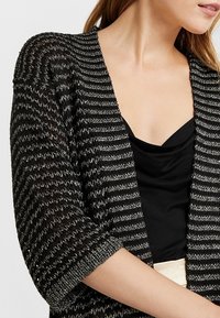 Öppen cardigan i svart och silverrandig stickad design, med korta dolmansärmar och en avslappnad passform. Mjuk textur med genomskinlig effekt.