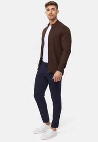 Braune Zip-Up-Strickjacke aus strukturiertem Material, getragen über einem weißen T-Shirt, kombiniert mit navyblauen Slim-Fit-Hosen und weißen Sneakers.