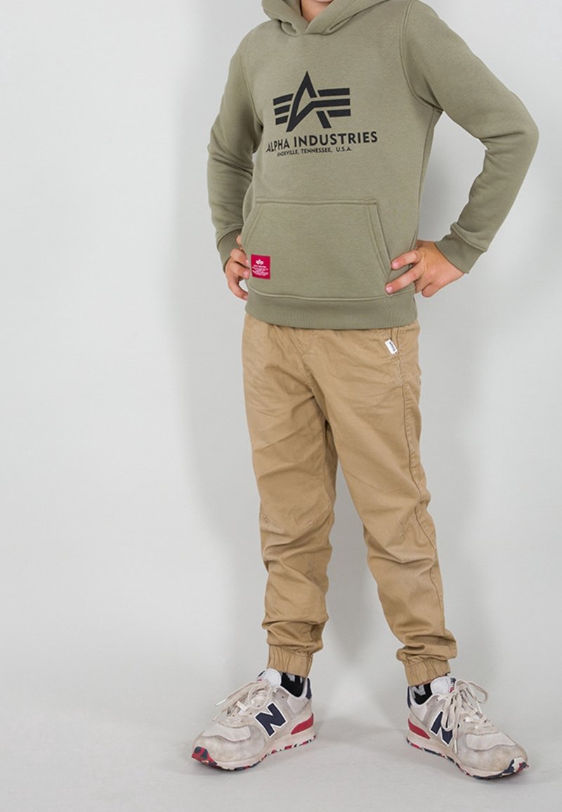 Sudadera verde oliva con logo negro, bolsillo frontal y puños acanalados, combinada con pantalones jogger beige y zapatillas blancas con acentos rojos.