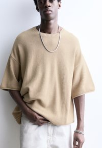 Béžový oversize pletený sveter s textúrovaným vzorom, okrúhlym výstrihom a krátkymi rukávmi. Strieborný retiazkový náhrdelník dopĺňa outfit.