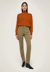 Spáleně oranžový rolák a olivově zelené přiléhavé džíny. Modelka má na sobě černé kotníkové boty. Hladká textura, na míru šitý střih, minimální detaily.