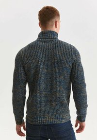 Pull en maille épaisse aux teintes bleues, marron et dorées. Col montant, motif texturé, manches ajustées et détail à ourlet côtelé.