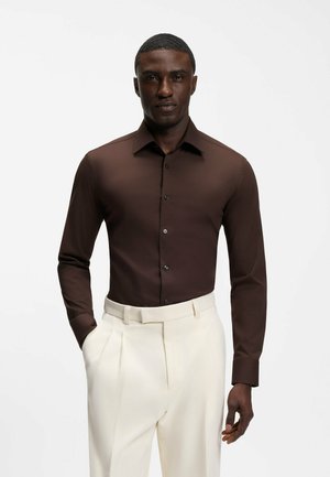 Uomo che indossa una camicia marrone aderente a maniche lunghe con bottoni, infilata in pantaloni crema a vita alta plissettati, con le mani parzialmente nelle tasche.