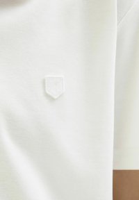 T-shirt blanc à manches courtes avec un logo en écusson brodé texturé sur la poitrine. Le tissu semble doux et respirant.