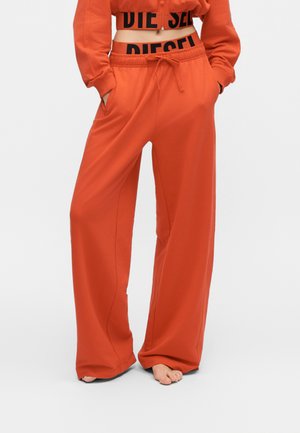 Personne portant un pantalon de survêtement orange ample avec les mains dans les poches et un haut court assorti montrant une ceinture noire Diesel.