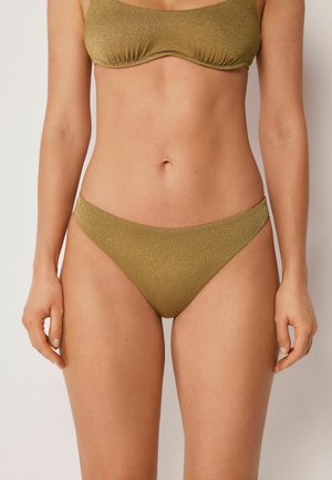 Frau trägt einen goldfarbenen, strukturierten Bikinihöschen und ein passendes Oberteil, steht mit entspannten Armen seitlich an ihrem Körper vor einem schlichten hellen Hintergrund.