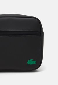 Schwarze Nylon-Kulturbeutel mit strukturiertem Oberflächen, oberem Reißverschluss und grünem Lacoste-Krokodil-Logo-Akzent. Rechteckige Form mit abgerundeten Kanten.