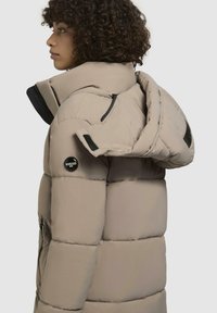 Jachetă puffer bej cu un guler înalt, textură matlasată, accente din metal negru și fermoare. Prezintă logo pe mânecă și pe glugă.