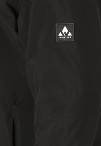 Schwarze Stoffhosen haben ein rechteckiges Patch mit weißem Berglogo und dem Text "WHISTLER". Glatte Textur mit minimaler Ausstattung.