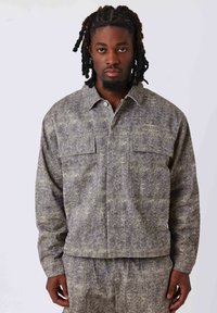 Veste à motifs gris et verts avec deux poches poitrine, une fermeture à boutons sur le devant et un tissu doux au toucher. Éclairée par un petit logo sur la poitrine.