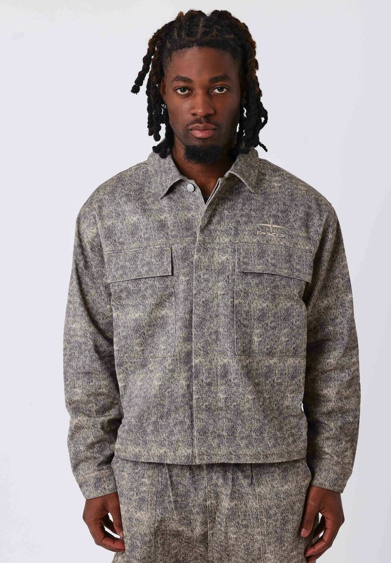 Veste à motifs gris et verts avec deux poches poitrine, une fermeture à boutons sur le devant et un tissu doux au toucher. Éclairée par un petit logo sur la poitrine.