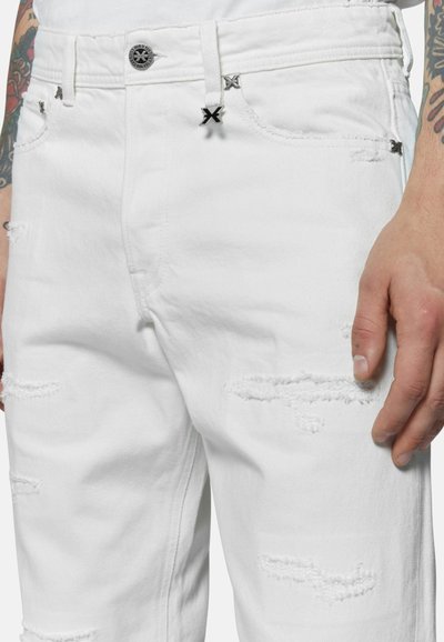 Jeans in denim bianco distressed con toppe sfilacciate e accenti in metallo scuro. Presentano una chiusura con zip e dettagli dei taschini.