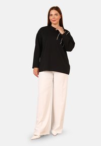 Top nero a maniche lunghe con scollo rotondo e dettagli decorati; abbinato a pantaloni bianchi a gamba larga e scarpe beige a punta.