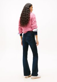 Woman with long wavy hair wearing a pink chunky sweater, dark blue flared jeans, and white sneakers, standing and facing away.