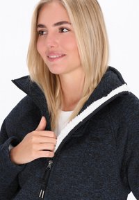 Giacca in pile di colore blu scuro con tessuto testurizzato, fodera in sherpa bianco sul colletto e chiusura a zip. Presenta una mezza zip e una vestibilità aderente.