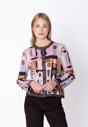 Młoda kobieta z prostymi blond włosami nosi kolorowy sweter na guziki w geometryczny wzór i czarne spodnie, stoi na białym tle.