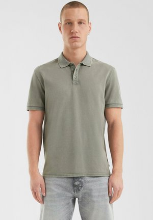 Polohemd aus einem gedämpften grünen Stoff, kurzärmelig, mit einer Dreiknopf-Leiste, gesticktem Kragen und Seitenkantenakzenten, kombiniert mit hellgrauen Jeans.