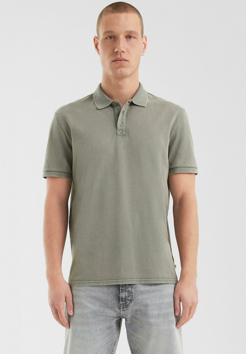 Poloshirt in gedempt groen stof, korte mouwen, drieknopenlijst, gestikt kraag en zijsplitaccenten, gestyled met lichtgrijze jeans.