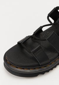 Sorte sandaler laget av teksturert lær med tre overremmer, elastiske snører og en tykk gummisåle med kontrasterende brune sømmer.