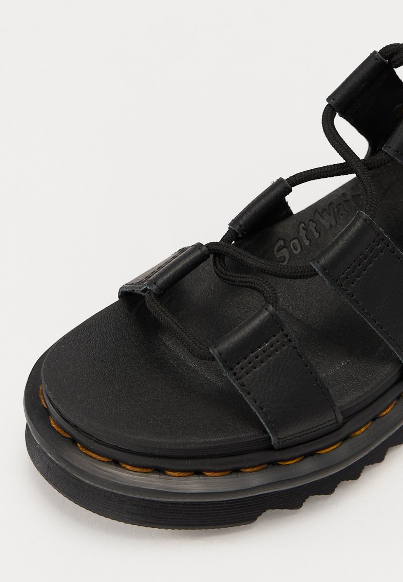 Black sandal made of textured leather with three upper straps, elastic laces, and a thick rubber sole featuring a contrasting brown stitch.