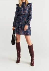 Robe bleu marine avec motif cachemire, col à volants, manches longues et jupe à volants. Portée avec des bottes hautes bordeaux et tenant un sac noir.