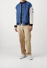 Kurtka bomberka z denimu z kontrastującymi białymi rękawami, zatrzaskami i detalem "HUG". Połączona z beżowymi spodniami i białymi trampkami.