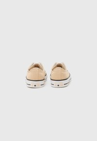 Beige kanvas sneakers med lavt design, hvit gummisåle og svarte detaljer. Teksturert overflate med synlige sømmer og logo på hælen.