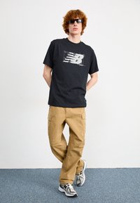 Svart t-shirt med kort ärm och vit New Balance-logo, tillsammans med beige cargobyxor. Modellen bär solglasögon och mönstrade sneakers.