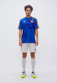 Young man standing in blue Real Madrid soccer jersey, white shorts, white socks, and cleats with neon yellow studs on plain background.