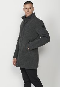 Abrigo de mezcla de lana gris con cuello alto, bolsillos laterales y un parche en el hombro detallado. Ajuste estructurado, textura suave y elementos de diseño clásicos.