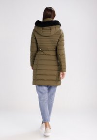 Žena odchází a má na sobě olivově zelený prošívaný kabát s černou kožešinovou kapucí, světle modré džíny a béžové tenisky.