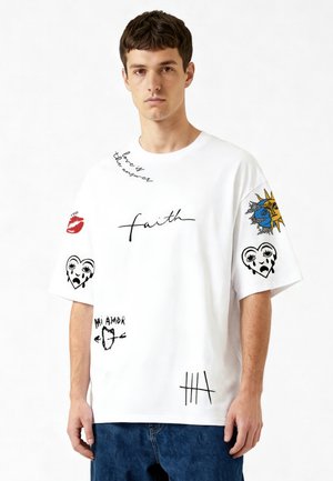 Jeune homme portant un T-shirt blanc oversize avec du texte et des graphismes, incluant « faith », soleil et lune, cœurs en pleurs, lèvres rouges et designs « Mi Amor ».