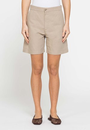 Persona che indossa pantaloncini beige sopra il ginocchio, top bianco e scarpe piatte intrecciate marroni, in piedi davanti a uno sfondo uniforme.