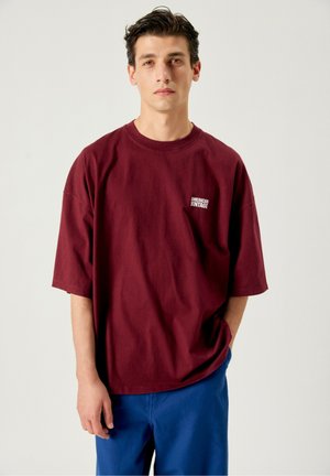 Jeune homme aux cheveux foncés porte un t-shirt bordeaux ample avec le logo "American Vintage" et un pantalon bleu, debout devant un fond uni.