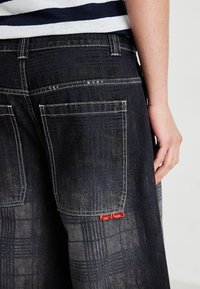 Persoană îmbrăcată cu blugi de denim în carouri închise, având buzunare mari la spate și o etichetă roșie mică lângă marginea buzunarului, mână sprijinită de partea corpului.