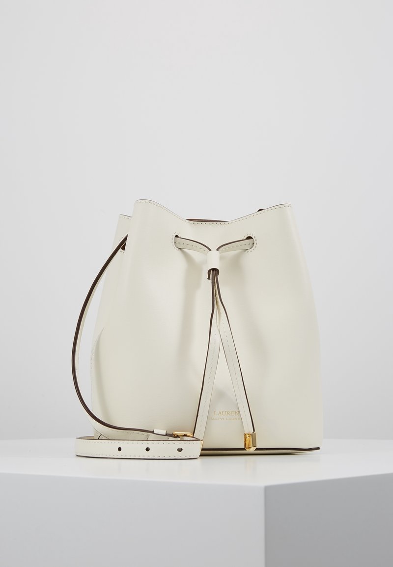 Krémová kožená kabela ve tvaru vědra s tahem na stáhnutí a nastavitelným popruhem, umístěná na bílé podstavě na bílém pozadí.