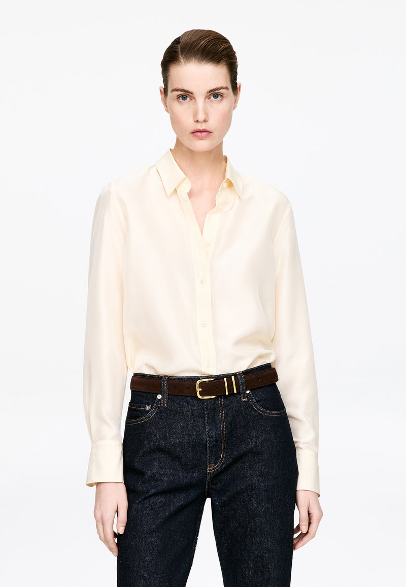Femme aux cheveux plaqués en arrière portant une chemise crème boutonnée rentrée dans un jean foncé avec une ceinture marron sur fond blanc.
