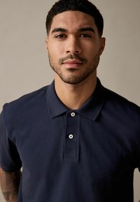Marineblå polo-shirt lavet af struktureret stof, med klassisk krave, korte ærmer og tre knapper ved blusen.