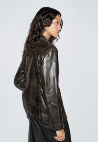 Vrouw met lang, krullend haar, gekleed in een donkerbruin leren jack en zwarte broek, zijwaarts gericht tegen een effen lichte achtergrond.